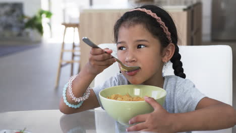 Biracial-girl-enjoys-a-bowl-of-cereal,-wearing-a-pink-headband-and-beaded-bracelets