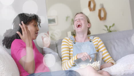 Animation-Von-Weißen-Lichtpunkten-über-Zwei-Unterschiedlichen-Teenager-Mädchen,-Die-Lachen-Und-Mit-Popcorn-Spielen