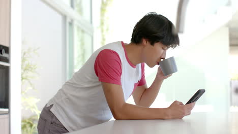 Teenage-Asian-boy-sips-coffee-and-checks-his-phone-at-home-in-the-kitchen