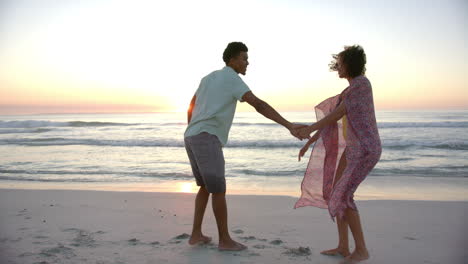 Biracial-couple-holds-hands-on-a-beach-at-sunset,-waves-gently-breaking-in-the-background