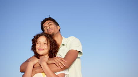 Biracial-couple-embraces-under-a-clear-blue-sky,-the-man-behind-the-woman-with-his-arms-around-her