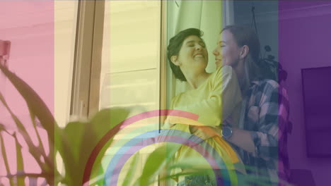 Animation-of-pride-rainbow-flag-and-rings-over-happy-caucasian-lesbian-couple-embracing-at-home