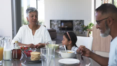 Biracial-grandparents-enjoy-breakfast-with-their-young-granddaughter,-surrounded-by-pancakes-and-fru