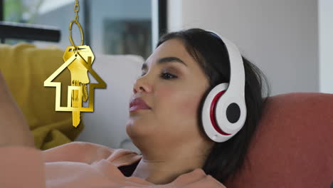 Animation-of-gold-house-key-and-key-fob-over-biracial-woman-using-headphones-at-home