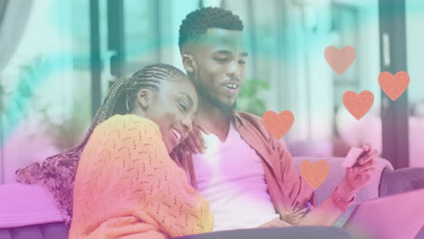 Animation-of-hearts-over-happy-diverse-couple-using-smartphone-at-home