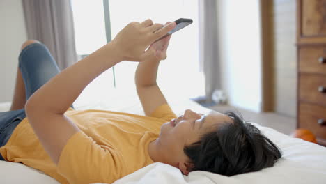 Teenage-Asian-boy-lies-on-a-bed-at-home,-browsing-his-smartphone