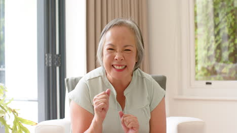Asian-senior-woman-with-grey-hair-smiles-with-closed-eyes,-sitting-indoors
