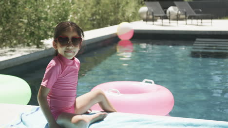 Biracial-girl-wearing-pink-swimwear-and-heart-shaped-sunglasses-sits-by-a-pool