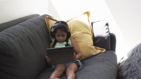 Biracial-girl-with-headphones-uses-a-tablet-on-a-gray-couch