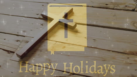Animation-of-happy-holidays-text-over-christian-cross-on-wooden-background