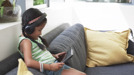 Biracial-girl-with-headphones-uses-a-tablet-on-a-couch