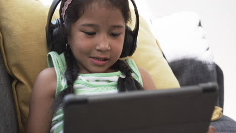 Biracial-girl-with-headphones-uses-a-tablet,-sitting-comfortably-on-a-yellow-cushion