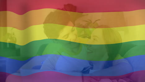 Animation-of-pride-rainbow-flag-over-happy-caucasian-lesbian-couple-using-laptop-in-bed