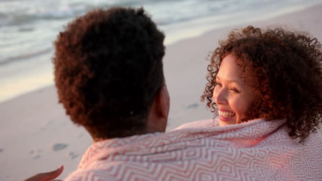 Biracial-couple-enjoys-a-sunset-at-the-beach,-the-woman-with-curly-hair-smiling-warmly