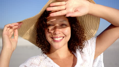 A-young-biracial-woman-with-curly-hair-smiles-on-a-sunny-beach,-shielding-her-eyes-with-her-hand
