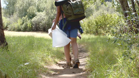 A-person-is-hiking-on-a-dirt-trail-carrying-a-large-backpack-and-a-white-container,-collecting-trash