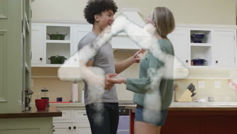 Animation-of-cloud-house-over-happy-diverse-couple-dancing-together-in-kitchen