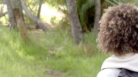 A-young-biracial-woman-with-curly-hair-gazes-into-a-lush-green-outdoor-setting-with-copy-space