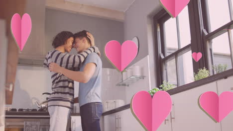 Animation-of-pink-hearts-over-happy-diverse-gay-male-couple-embracing-in-kitchen