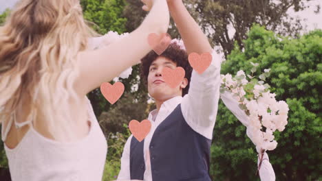 Animation-of-pink-hearts-over-happy-diverse-couple-dancing-in-garden-on-wedding-day