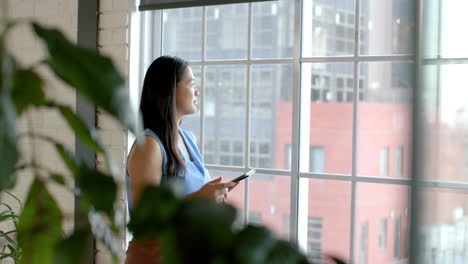 Asian-businesswoman-stands-by-a-window-in-an-office-setting,-holding-a-tablet,-with-copy-space