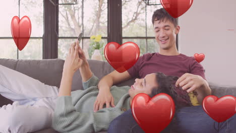 Animation-of-red-hearts-over-happy-diverse-gay-male-couple-relaxing-on-couch-using-smartphone