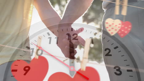 Animation-of-moving-clock-and-hearts-over-caucasian-couple-holding-hands