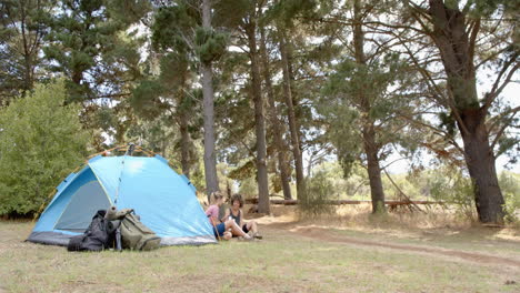Two-women-relax-beside-a-blue-tent-in-a-wooded-campsite-with-copy-space