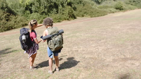Two-women-with-backpacks-are-hiking-in-a-sunny,-grassy-field-with-copy-space