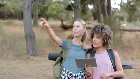 Young-Caucasian-woman-points-into-the-distance-as-a-young-biracial-woman-looks-on,-holding-a-tablet