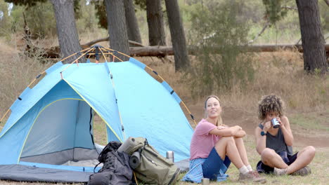 Young-Caucasian-woman-and-biracial-woman-relax-beside-a-blue-tent-in-the-woods