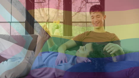 Animation-of-rainbow-flag-over-happy-diverse-gay-male-couple-relaxing-on-couch-using-smartphone