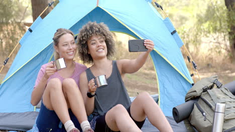 Two-women-sit-inside-a-tent,-one-holding-a-mug-and-the-other-taking-a-selfie