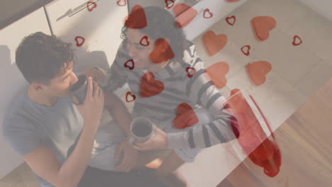 Animation-Von-Roten-Herzen-über-Einem-Glücklichen-Schwulen-Paar,-Das-Auf-Dem-Küchenboden-Sitzt-Und-Kaffee-Trinkt
