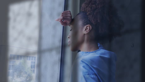 Animation-of-rain-on-window-over-unhappy-african-american-female-doctor-looking-out-of-window