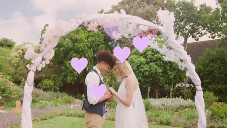 Animation-of-lilac-hearts-and-happy-diverse-couple-holding-hands-under-wedding-arch-in-garden