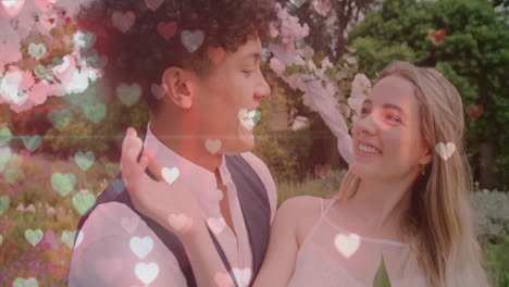 Animation-of-glowing-hearts-over-happy-diverse-couple-smiling-in-garden-on-wedding-day
