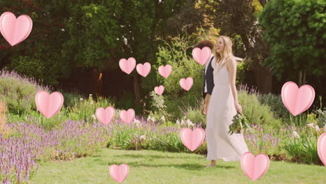 Animation-of-pink-hearts-over-happy-diverse-couple-walking-in-sunny-garden-on-wedding-day