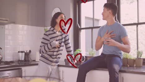Animation-of-red-hearts-over-happy-diverse-gay-male-couple-talking-and-laughing-in-kitchen