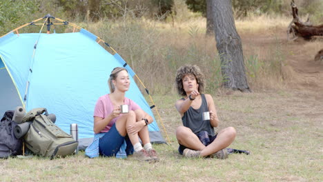 Two-women-relax-by-a-tent,-one-holding-a-mug-and-the-other-using-binoculars