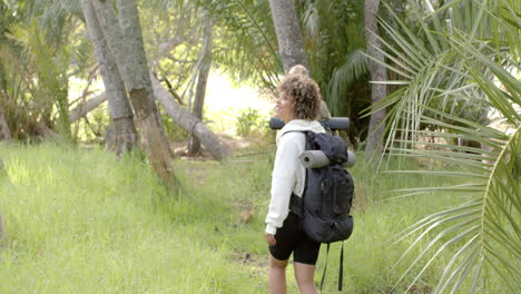 Young-biracial-woman-with-curly-hair-carries-a-backpack-in-a-lush-forest-with-copy-space