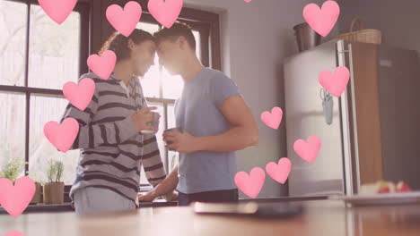 Animation-of-pink-hearts-over-happy-diverse-gay-male-couple-touching-heads,-having-coffee-in-kitchen
