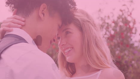 Animation-of-hearts-and-pink-tint-over-happy-diverse-couple-embracing-at-outdoor-wedding