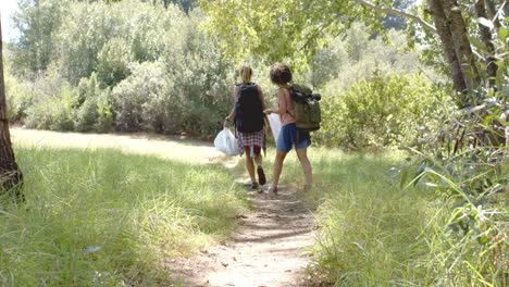 Two-women-walk-along-a-narrow-trail-in-a-lush,-green-forest-setting,-collecting-trash