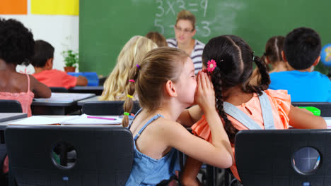 Schoolgirl-whispering-into-her-friend-s-ear-in-classroom