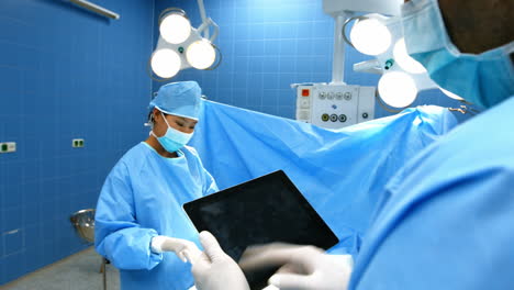Cirujano-Usando-Tableta-Digital-Mientras-Un-Compañero-De-Trabajo-Opera-Al-Paciente-En-El-Quirófano