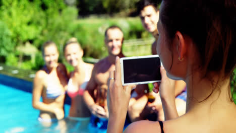 Smiling-woman-clicking-photos-of-friends-from-mobile-phone-near-poolside