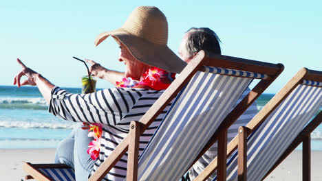 Älteres-Paar-Sitzt-Auf-Sonnenliege-Und-Trinkt-Cocktail-Am-Strand