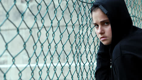 Portrait-of-sad-schoolgirl-in-hooded-standing-near-fence