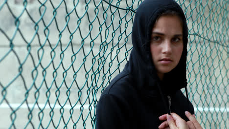 Portrait-of-sad-schoolgirl-in-hooded-leaning-against-fence